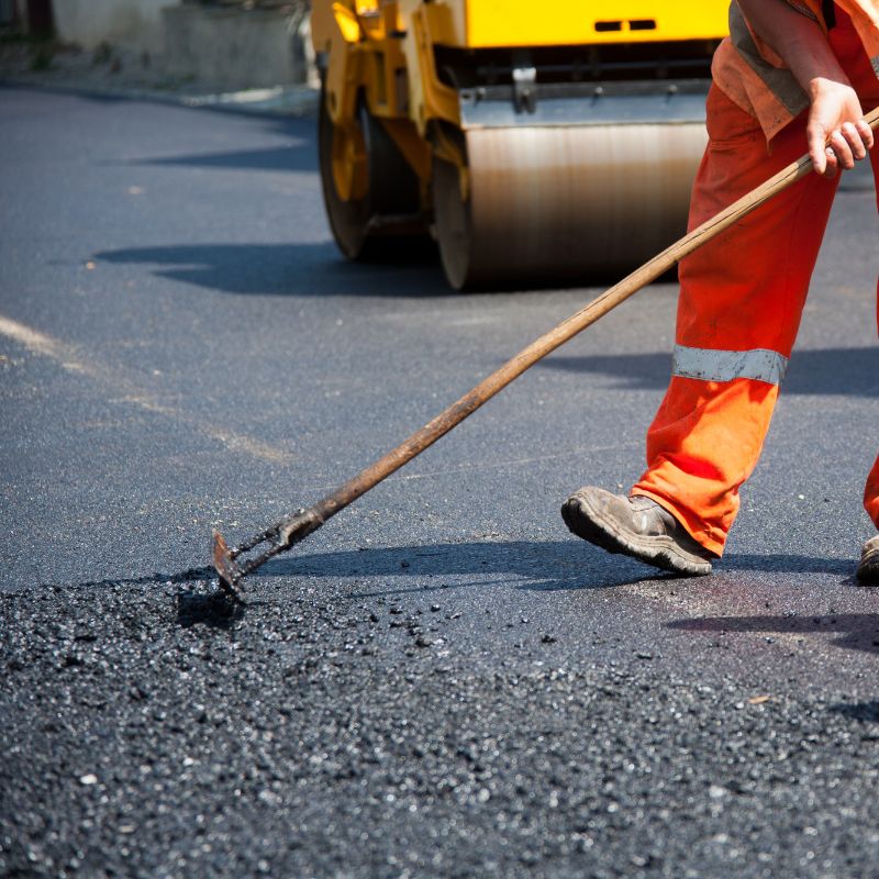 Worker in orange uniform smoothing fresh asphalt with a rake, expertly handling crack filling duties, as a yellow steamroller stands ready in the background.