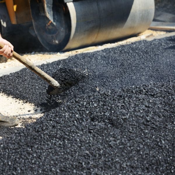 Construction worker using a shovel to spread asphalt on a road with a steamroller in the background.