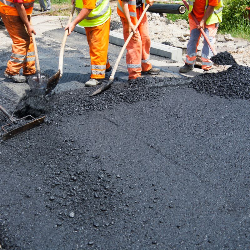 Workers in orange uniforms meticulously spreading asphalt to repair potholes on the road with shovels and rakes.