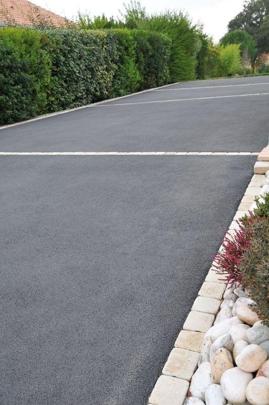 A smooth asphalt driveway with white lines, bordered by bushes and a row of decorative stones in the foreground.