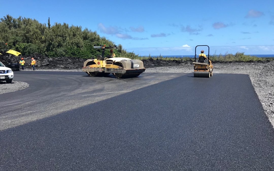 Construction workers paving a road with machinery, surrounded by greenery and a view of the ocean.