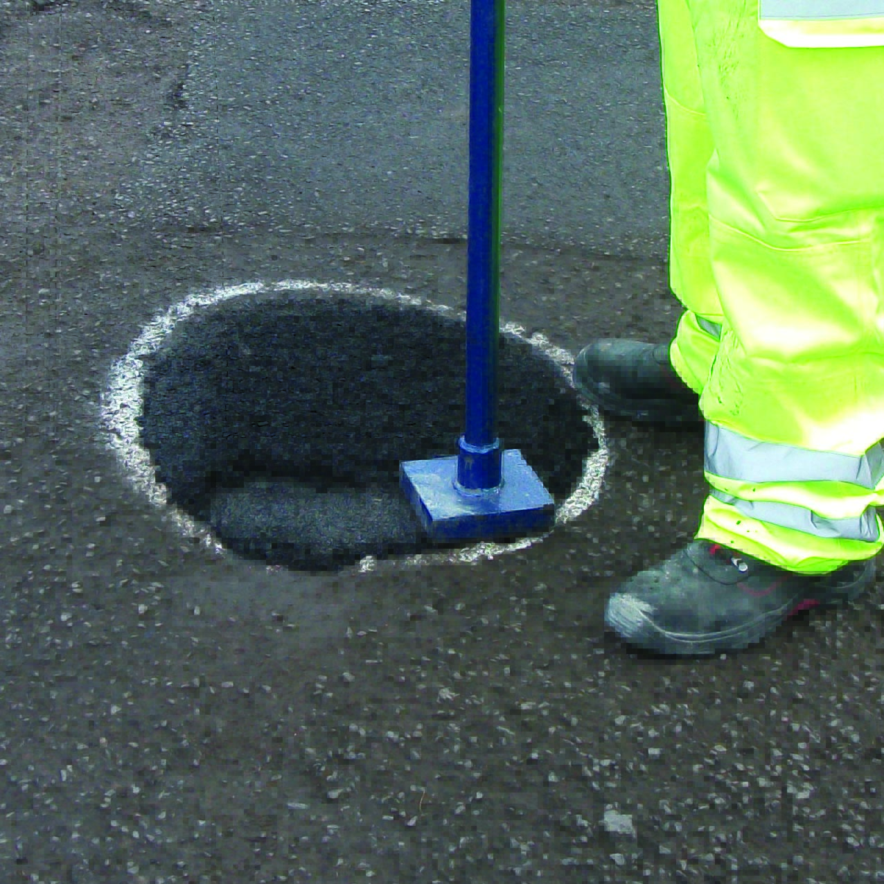 A worker in neon safety gear stands proudly next to a completed pothole repair on the asphalt road.