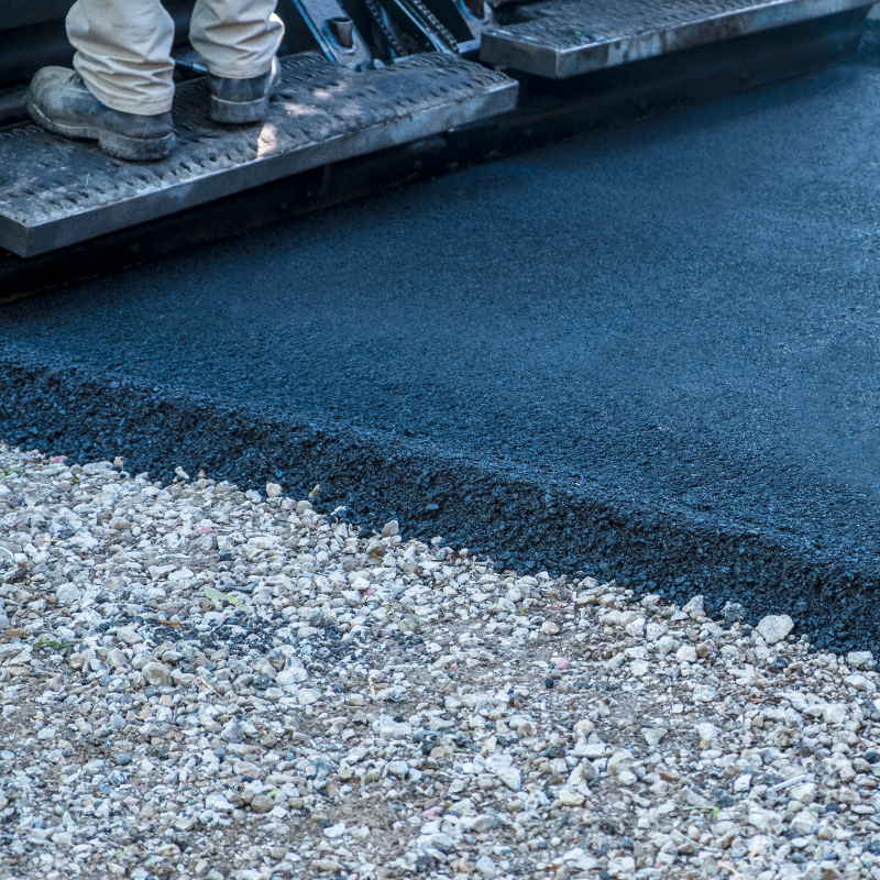 Close-up of workers paving a road with fresh asphalt, showing their legs and paving machinery.