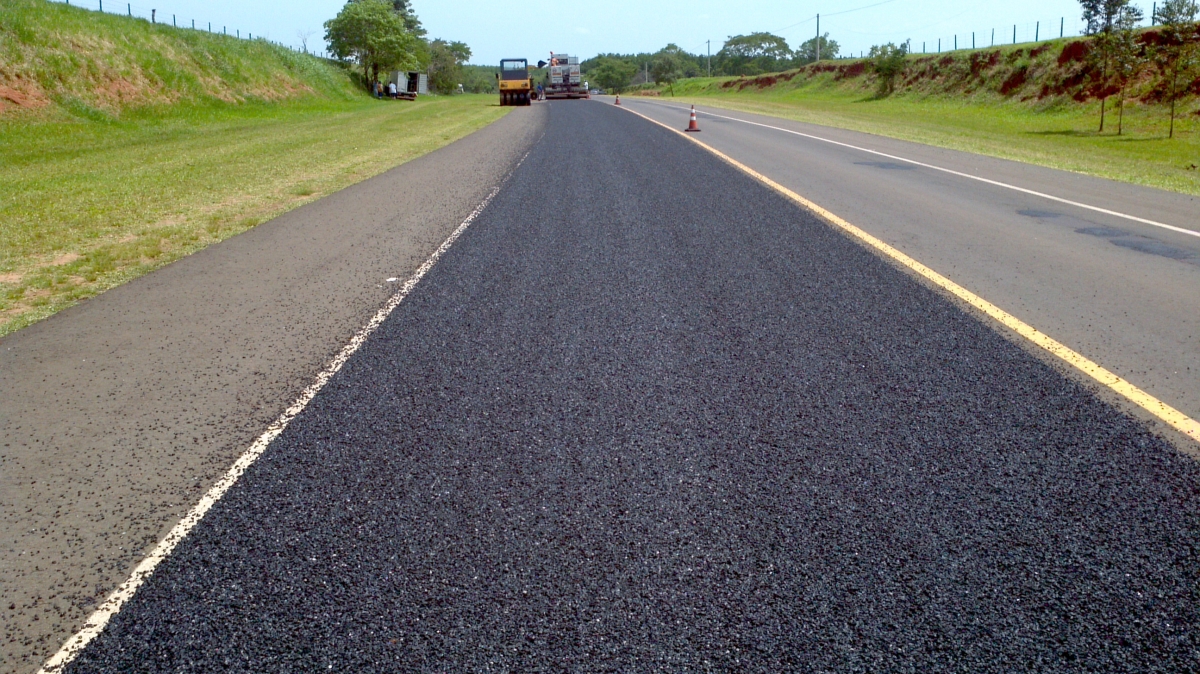 Freshly paved road with construction vehicles in the distance, surrounded by green grass and trees.