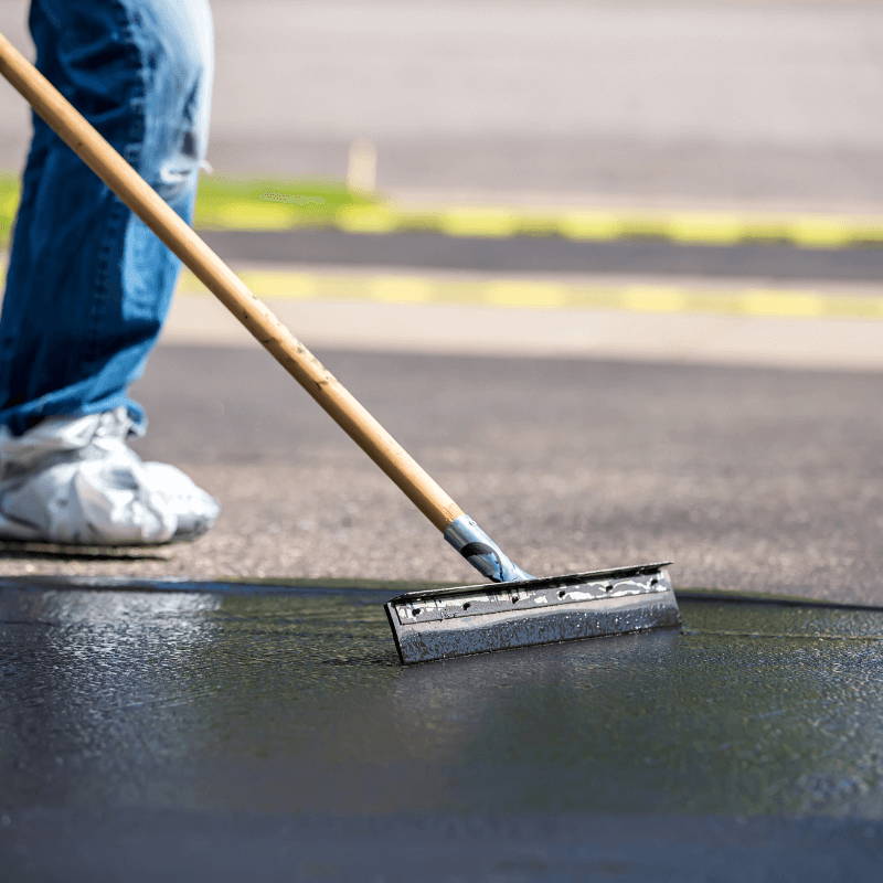 Person spreading asphalt sealer on a driveway with a squeegee tool, wearing jeans and sneakers.