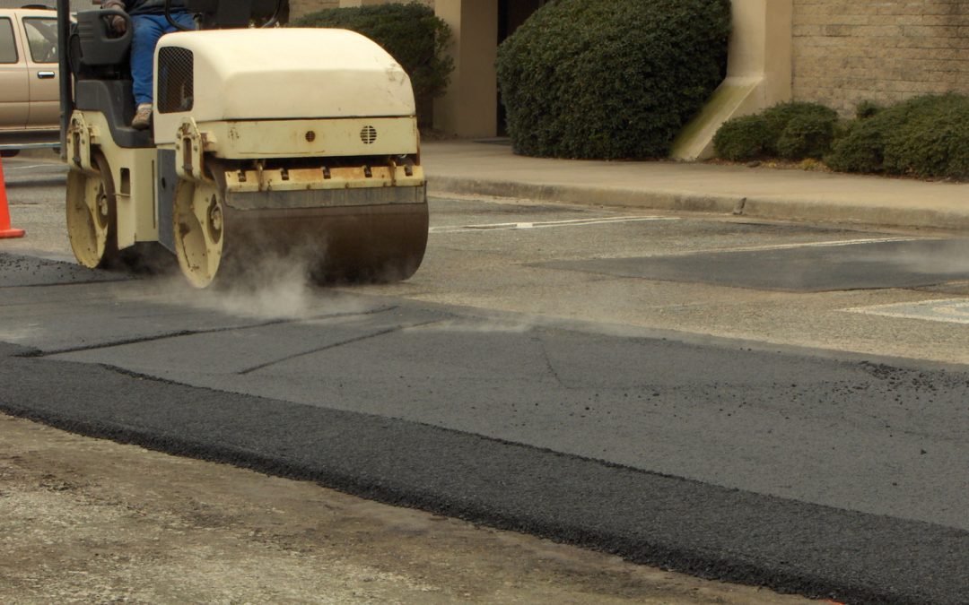 A worker operates a steamroller on freshly laid asphalt in a parking lot beside a concrete building.