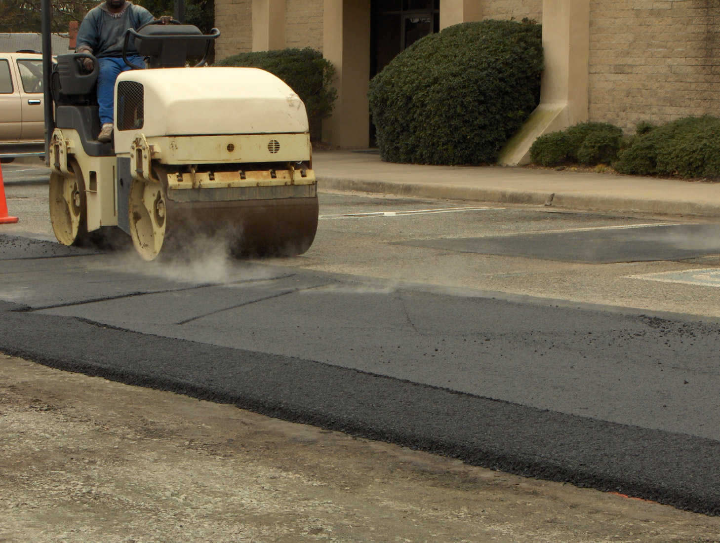 A worker operates a steamroller on freshly laid asphalt in a parking lot beside a concrete building.